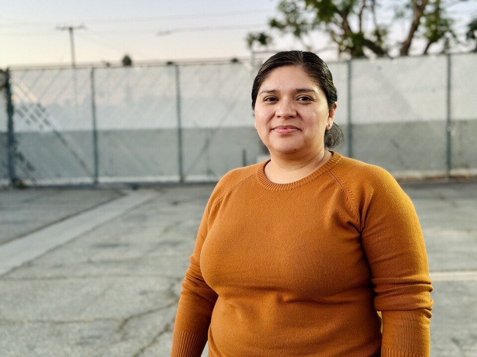 A medium-light skin-toned woman in a mustard-colored sweater stands outdoors, smiling against a backdrop of chain link fencing and foliage.