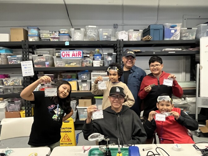 A group of kids show off their home-made ham radios. They're sitting and standing in front of shelves crammed with different kinds of gear.