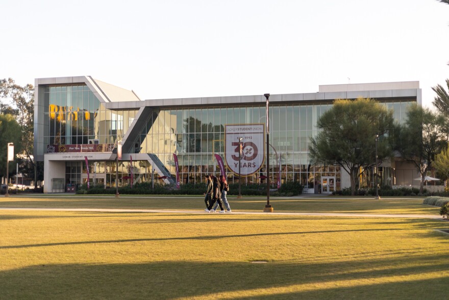 Three students on a grassy field can be seen walking along a concrete walkway, behind the grassy field, there's a glass and concrete modern building lined with green bushes. On the glass windows there's a large white sign that reads in red letters: "Locker Student Union Est. 1992. 30 Years."