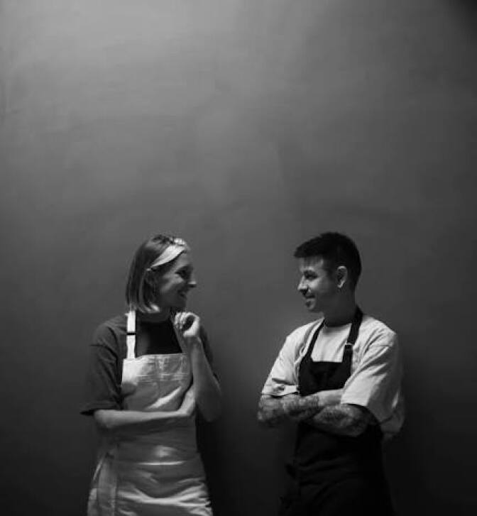 Black-and-white portrait of two ALTO chefs standing against a plain gray backdrop, facing each other in profile. The woman on the left wears a white apron over a T-shirt, her hair pulled back, one hand resting thoughtfully at her chin as she smiles. The man on the right wears a dark apron over a light T-shirt, arms crossed, a tattoo visible on his forearm, smiling back at her.