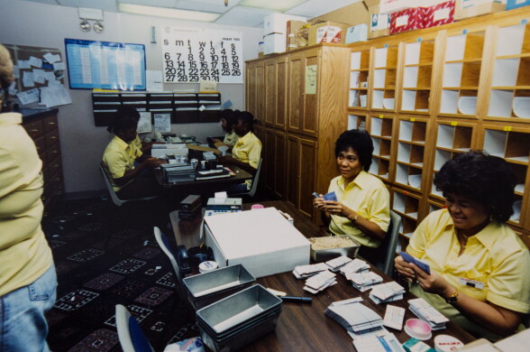 A group of employees sit in a backroom with stacks of cards and bins on the table. They all appear to be women with medium to dark skin tones, wearing yellow shirts.
