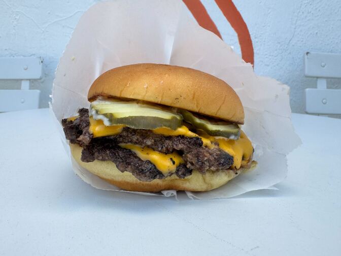 A close-up of a cheeseburger on a white surface with white to-go paper loosely wrapped around the burger itself. In the background, a white wall with two white chairs is partially shown. 