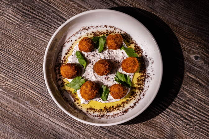 Seven golden fried olives arranged in a circle atop a swirl of white sauce, garnished with herbs and spice, on a white plate.