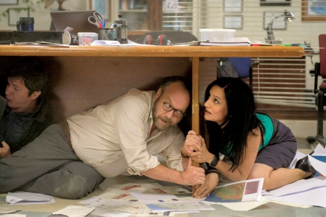 In a scene from the film 'San Andreas' Paul Giamatti and Archie Panjabi cling to the leg of a desk while crouched on the floor where papers are scattered.