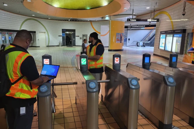 The interior of an underground public transit station. Two men in green and orange high-visibility vests and long pants are standing around the silver metal turnstile gates. The man on the left is working on a laptop computer at the same time. The station is largely empty and well-lit, except for one woman walking in the background. 