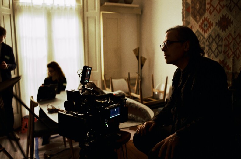 Un hombre con gafas a la sombra junto a la cámara. Un tapiz de pared aparece detrás de él. Hay personas al otro lado de la habitación cerca de una ventana cubierta con cortinas transparentes.