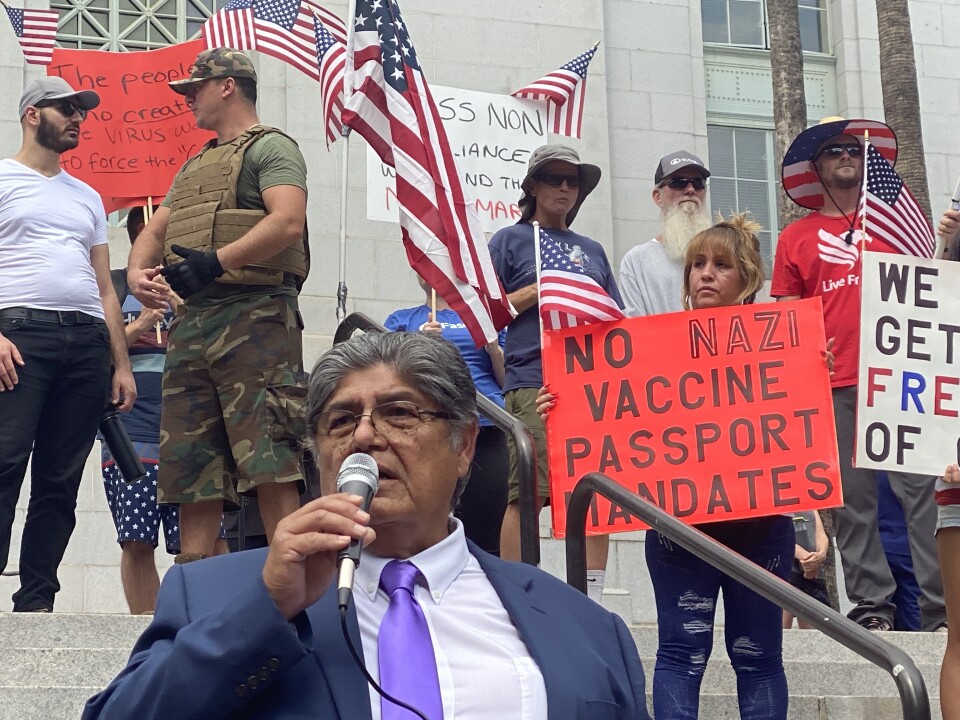 At the anti-vaccination rally in downtown LA on Aug. 14, 2021, a man in a blue suit with a purple tie, white shirt and salt-and-pepper hair is in the foreground, holding a microphone as he addresses the crowd. He's at the bottom of the steps to LA City Hall. Behind him to the right a woman holds a red sign that says in all capital letters, "No Nazi vaccine passport mandates." There are five men behind her. She has a small American flag attached to her sign; there are six other small American flags attached to signs behind her. Their messages are obscured. At the top of the steps a man in military fatigues with a camouflage cap, flak jacket and a black glove on his left hand is talking to a bearded man in dark pants, white t-shirt and tan baseball cap.