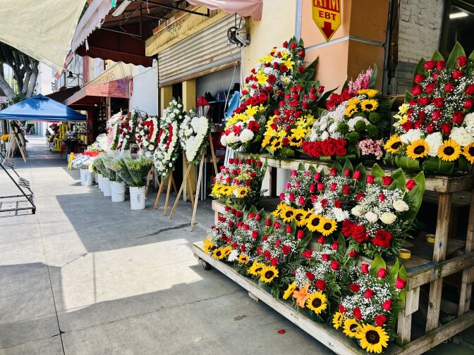 Different flower arrangements, in red white and yellow colors, sit on shelves in front of two stores.