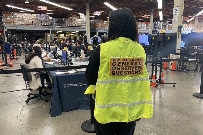 In the foreground, a woman with her back turned wears a bright yellow vest with a sign that reads "Ask me about general observer questions." In the background, a row of people sit in front of computers. 