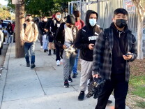 Male high school students in hoodies and joggers stand in line on a sidewalk outside their high school.