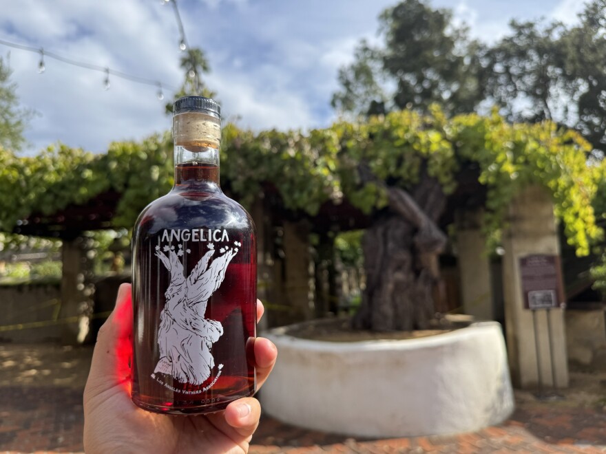 A hand holds a bottle of deep red Angelica wine with a white grapevine illustration on the label, photographed outdoors with Mission San Gabriel's historic grapevine arbor visible in the blurred background.