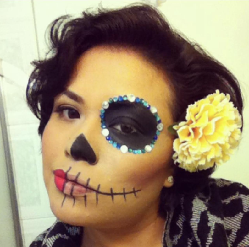 Flor in calavera makeup has black around her left eye, and a rime of jewels. Her mouth has cross hatches on the left and she wears a yellow flower behind her ear.