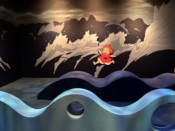 An interior room with dark waves painted on the walls, with an animated young girl poking out from on top of the waves. The young girl has red hair and is wearing a red dress with white pants. Two lighter blue foam seats are placed in the front of the wall, curved to resemble real ocean waves.