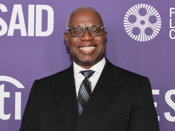A man with dark brown skin wearing eyeglasses and a black suit smiles broadly in front of a purple background