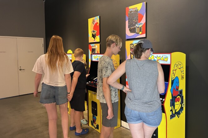 Three old-school PAC-MAN video arcade games are lined up against a black wall, with several people standing around them, playing. 