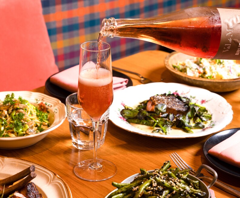 A bottle of bubbly rose wine is being poured into a flute on a table. Various dishes are also present, including asparagus, a salad and a steak on greens.