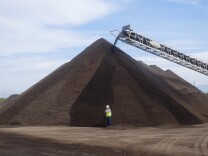A huge pile of dark brown compost in a dirt lot dwarfs a man wearing a white hard hat and neon safety vest.