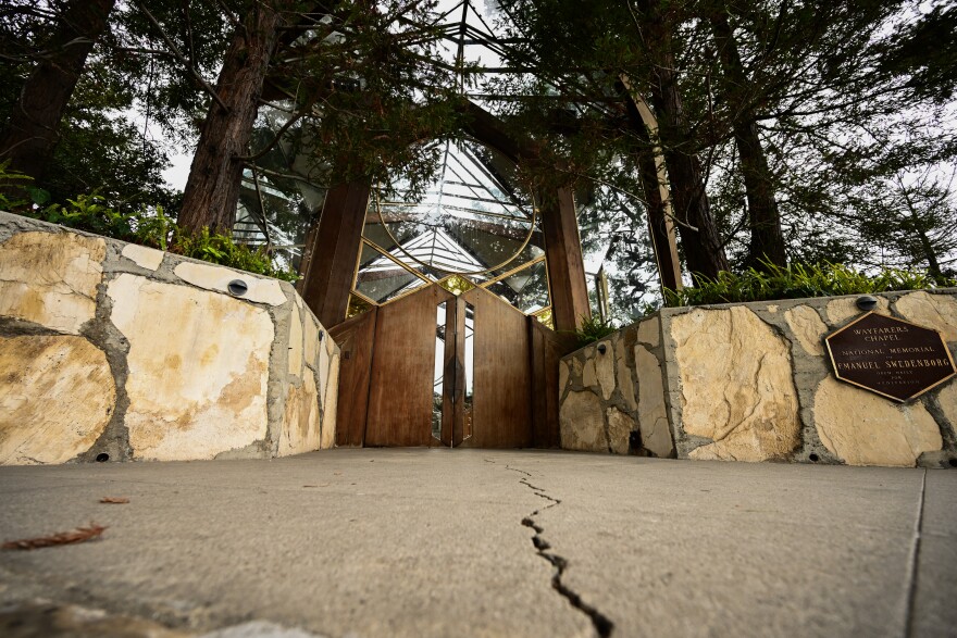 A large crack is visible in the walkway leading up to glass and wood doors of a chapel that is largely glass and wood beams.