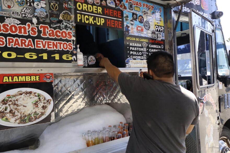 A man with medium skin tone wearing a gray t-shirt is holding a phone to his ear while ordering from a food truck.