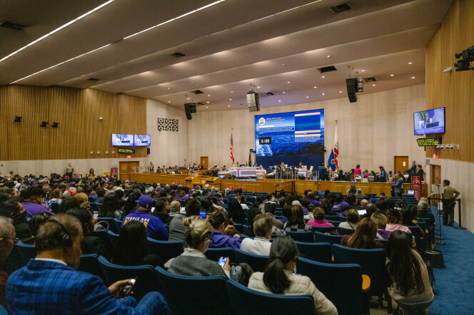 A large auditorium where a large crowd sits in blue chairs facing a dais with a large screen above. 