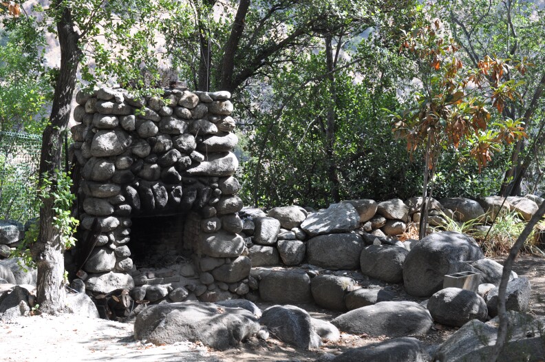 A large, round fire pit made entirely of grey stones. It is outdoors and surrounded with trees. 
