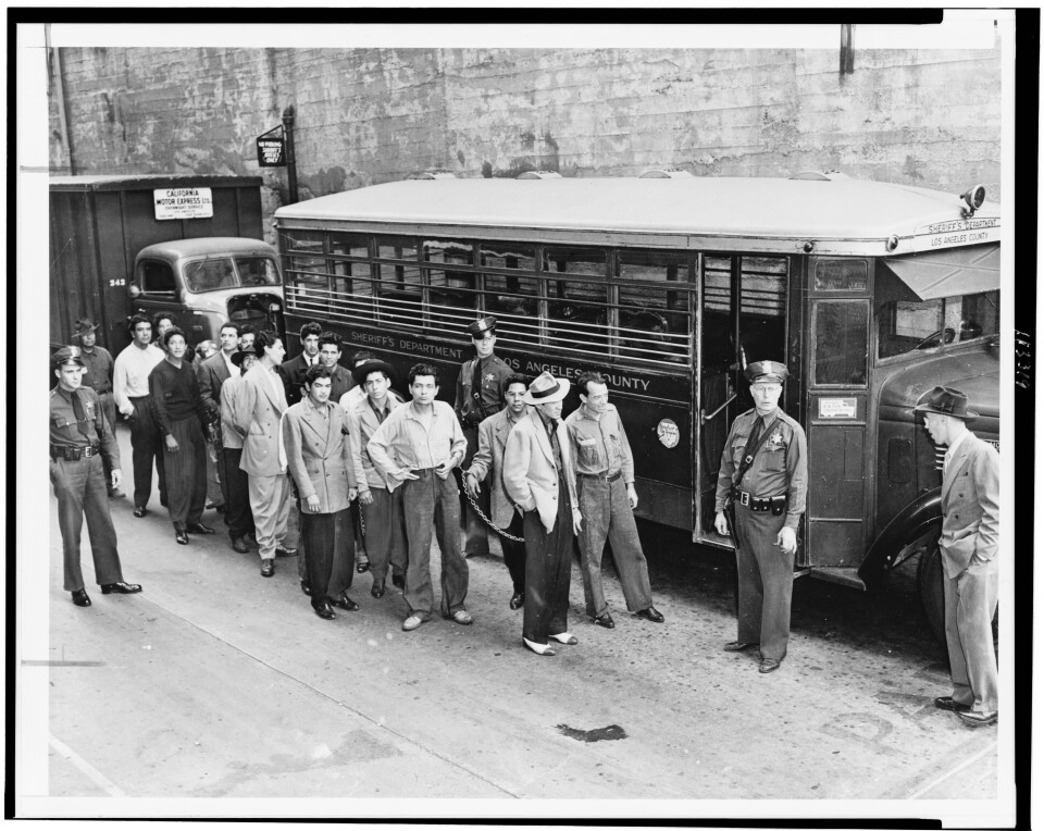 A vintage black-and-white photo shows a large group of men in baggy zoot suits, waiting to be loaded by officers onto a bus parked next to them.