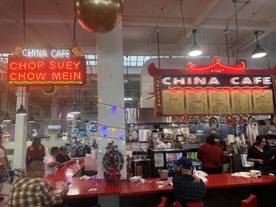 The neon signs above this particular food stall proclaim "China Cafe" and "Chop Suey Chow Mein," and a menu busy with Chinese food offerings hangs overhead: There are several people working behind the red counter, while several diners sit at the very same counter, enjoying their meals.  
