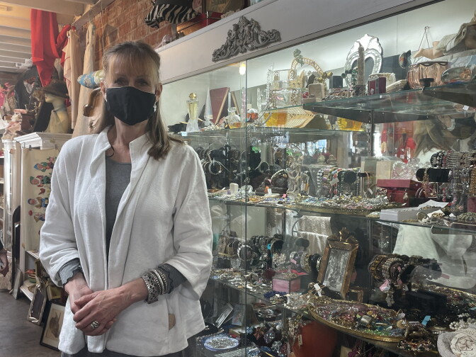 a woman stands in a store with all sorts of tchotchkes and other items cramming the glass shelves. she wears a black face mask