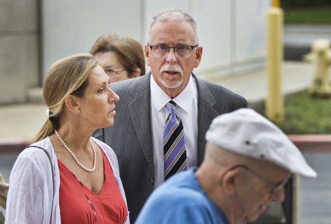 A man with light skin tone, short gray hair, and a gray goatee, wearing glasses and a charcoal suit, looks at the camera as two people, standing in the foreground and slightly out of focus, look to their right.