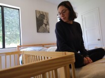A woman with medium-tone skin and glasses looks down on a wood crib while sitting on a bed. The photo of a woman hangs on a white wall between windows.