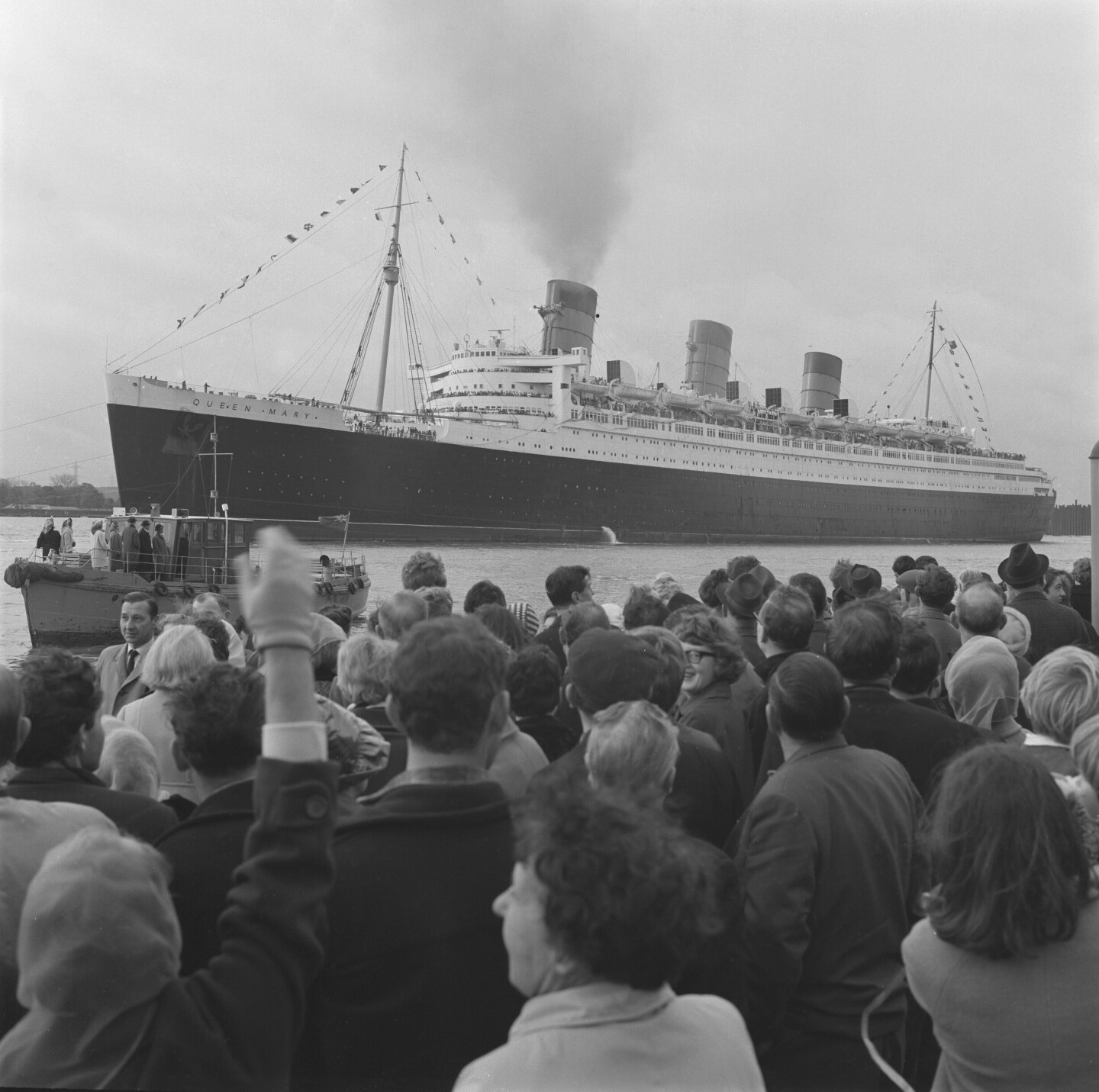 In a black and white image the Queen Mary departs the docks in Southampton, England bound for Long Beach with a crowd waving goodbye.