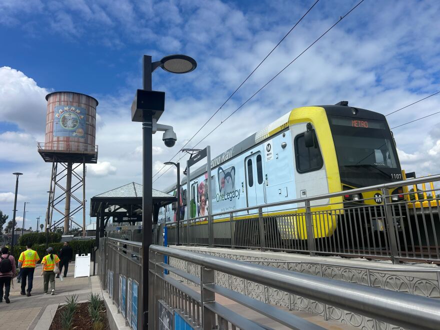 A light rail car is on the tracks at a station. A water tower stands in the distance with people nearby walking along a sidewalk.