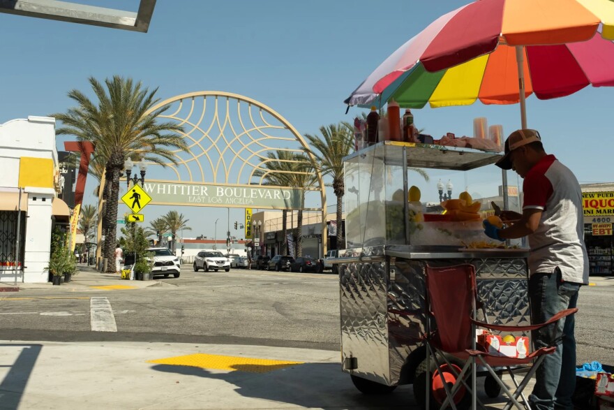 A street vendor stands under a rainbow umbrella peeling fruit in front of a cart. In the distance is an arch with a sign that reads "Whittier Blvd. East Los Angeles."