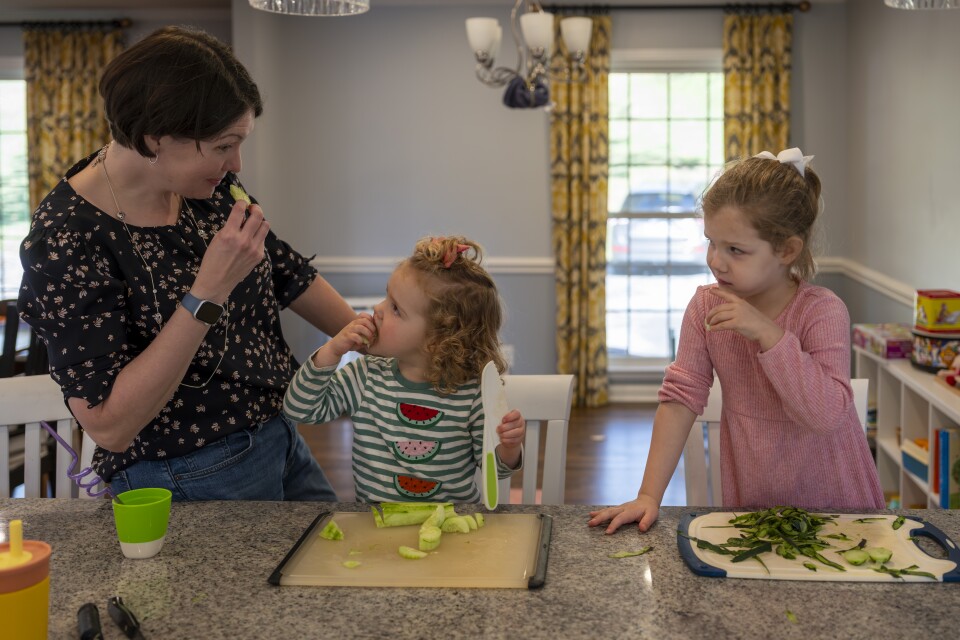 A woman with light skin tone, wearing a black shirt with floral print, eats a slice of cucumber facing two children sitting and standing on chairs with peeled and cut cucumbers on cutting boards on a counter.