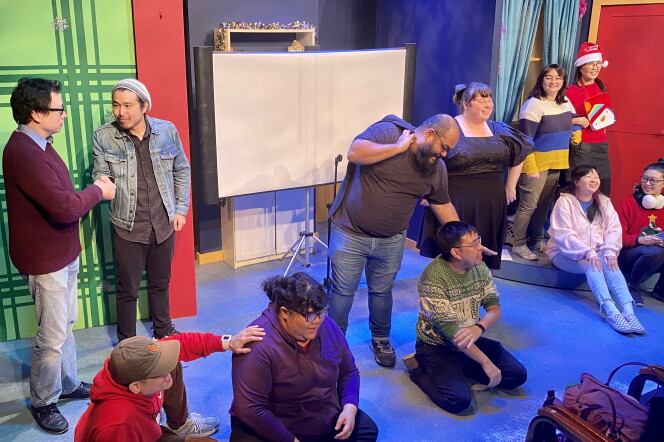 Eleven Asian people are spread out across a small, colorful stage, in various poses, some standing, some sitting on the ground.