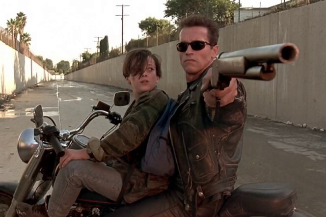 A young boy and a man wearing sunglasses and a leather jacket sit on a motorcycle, turned sideways in a flood-control channel. The man is pointing a rifle at something behind them while the boy looks at the man's face.