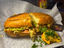 A toasted Guatemalan torta sandwich filled with shredded meat, lettuce, tomatoes, and guacamole, served on parchment paper with a side garnish of parsley and tomato. A small toothpick flag with “Muchá! Comida Chapina” is placed beside it.