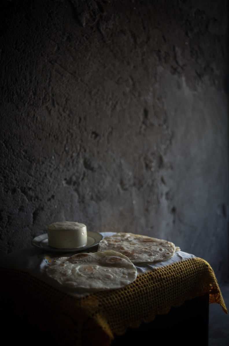 A semi-lit color photograph shows a small table with a yellow tablecloth holding tortillas and a round of cheese, set against a darkened wall.