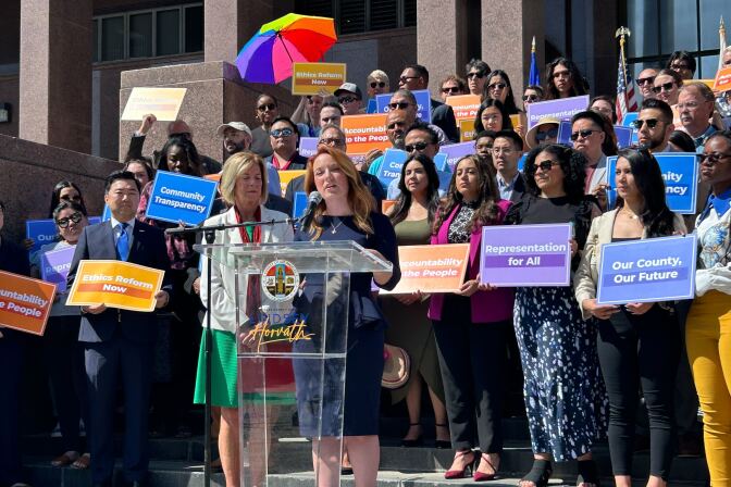 Two women stand at a podium with several people behind them holding signs of various colors. One woman at the podium has chin-length blonde hair and is wearing a green dress with a white jacket. The other woman has auburn hair that reaches past her shoulders. She is wearing a dark blue dress.