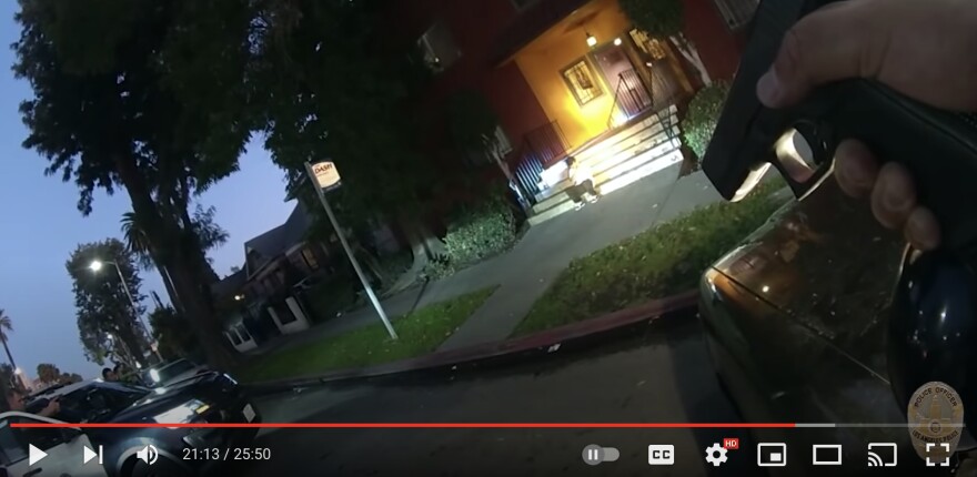 A screen shot from the YouTube upload of LAPD body-worn camera video of the fatal Margarito López shooting. In the foreground, an LAPD officer can be seen standing behind his car and pointing a firearm at López. López sits on the bottom step of an entryway staircase outside of a home. There is a spotlight pointed on López. It is dusk.