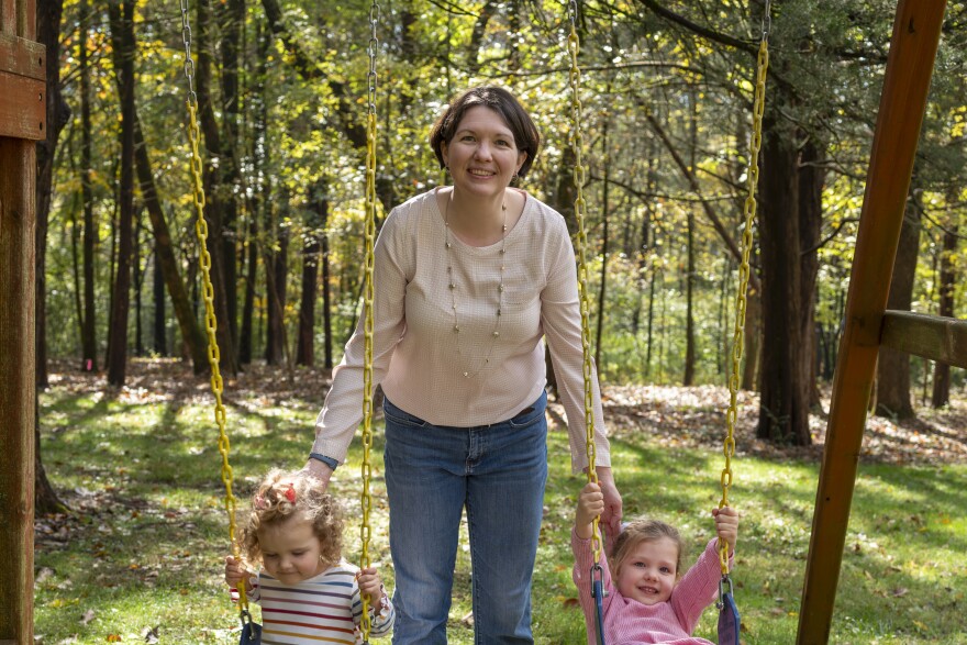 A woman with light skin tone, wearing a cream-colored sweater and jeans, pushes two small children on a swing set with trees in the background.