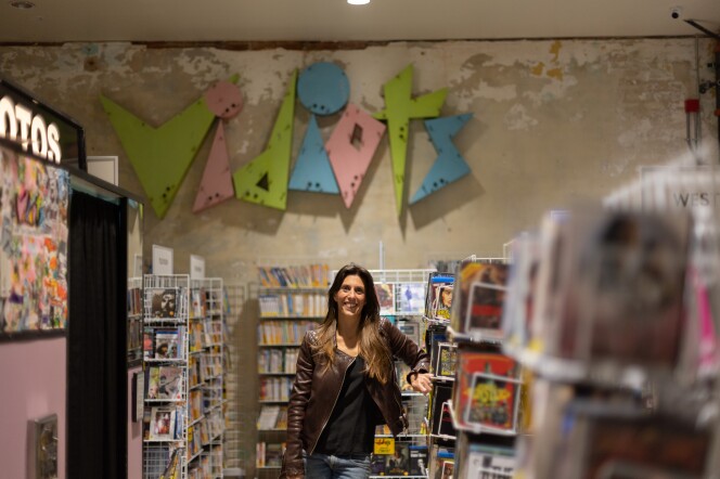 A feminine presenting person with light skin tone and light brown hair wearing a brown leather jacket and jeans stands leaning on a stand full of magazines. Behind them on the wall is a colorful sign that reads "Vidiots."