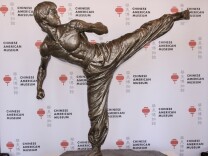 A fiberglass statue of martial arts legend Bruce Lee is set against a backdrop printed with a red lantern logo for the Chinese American Museum.