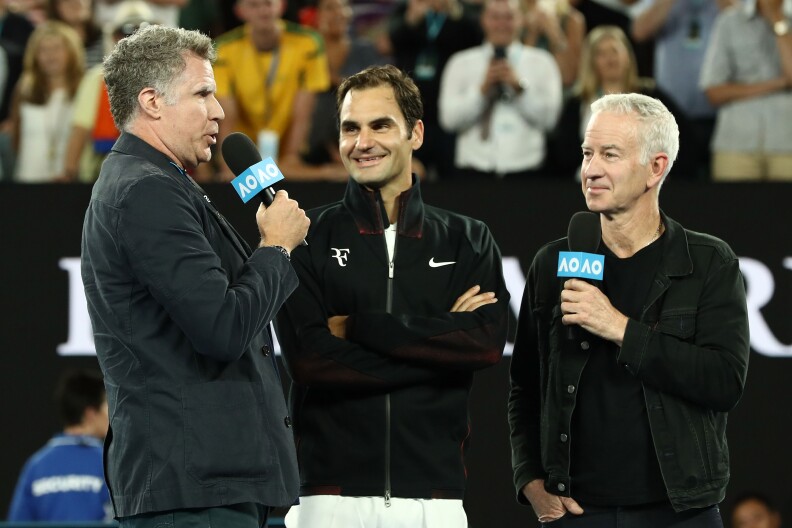 Will Ferrell holds a mic on the left; to his right is Roger Federer arms folded in a zip-up sweater, then on the far right is John McEnroe holding a microphone. Fans in the stands are seen in the background.
