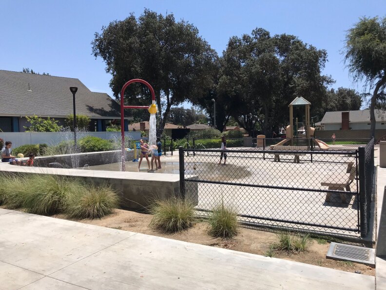 A view of the park from the sidewalk. There are children playing with a sprinkler installation behind a fence.