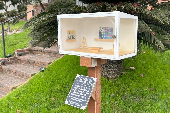 A white box with two transparent walls that showcase small works of art, including a ceramic nose and drawings. The box, located in a home's front yard, is accompanied with a sign that reads "Museum of Free Art"