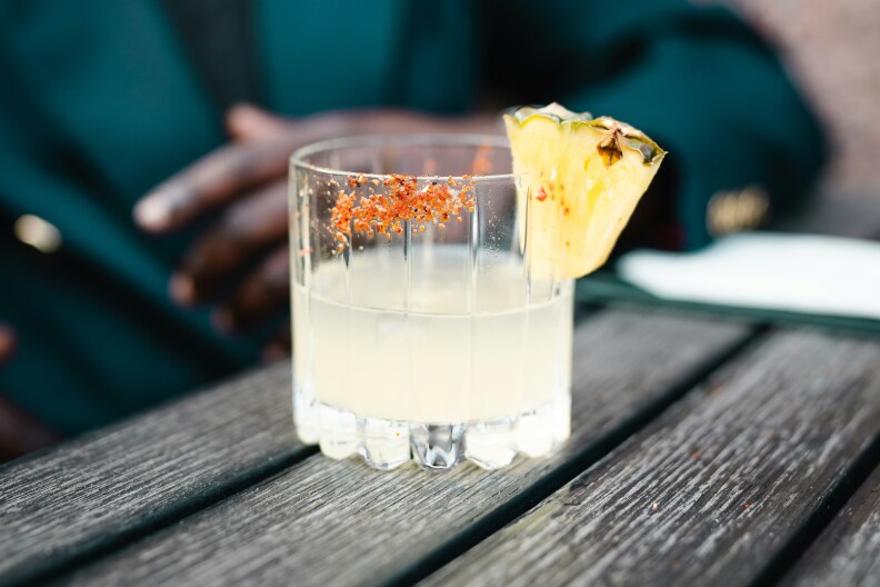Image of a glass of Tequila in the center of the image and only thing in focus. The drink is sitting on top of a wooden table with a man behind the drink, out of foucs.