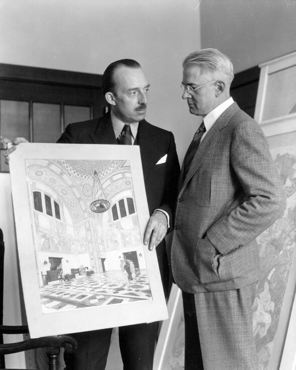 A black and white archival look at two white men standing while facing each other. The man on the left, who has a mustache and suit, is holding up an illustration of the mural idea, which show the library's rotunda ceiling. The man on the right, who has a suit and glasses on, is looking at the artwork.