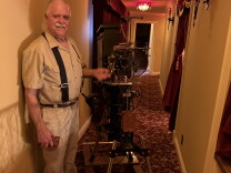 An older man with a gray mustache wearing a tan shirt, white pants and suspenders is handcranking an antique movie projector