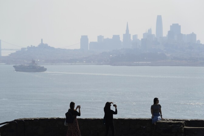 Three people are pictured in silhouette, standing at the edge of a bay taking photos. In the distance is the San Francisco skyline, shrouded in smoke.